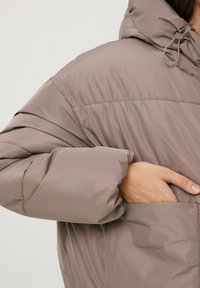 Giacca a vento imbottita in un morbido colore taupe, con cappuccio, polsini elastici e una tasca frontale con una texture liscia e lucida.