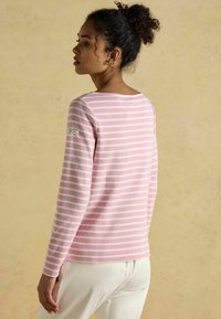 Svetloružový pruhovaný top s dlhými rukávmi a okrúhlym výstrihom, s bielymi pruhmi. Obsahuje malú nášivku s logom na ľavnom rukáve. Štruktúrovaná tkanina.