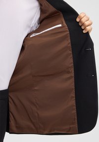 Giacca nera su misura con foderatura in satin marrone. L'interno presenta una tasca con zip e cuciture dettagliate lungo i bordi.