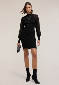 Schwarz geripptes Kleid mit transparenten Puffärmeln, figurbetonter Silhouette, knielang, kombiniert mit einer strukturierten Clutch und Stiletto-Stiefeln.