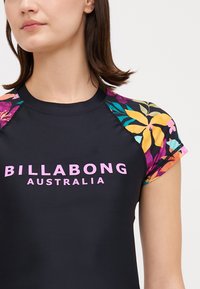 Black rash guard with colorful floral-patterned short sleeves. Features "BILLABONG AUSTRALIA" in bold pink text across the front.