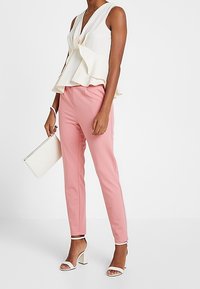 Valkoinen peplum-pusero V-pääntiellä, yhdistetty vaaleanpunaisiin räätälöityihin housuihin. Valkoinen laukku ja remmillä varustetut korkokengät täydentävät asun. Tasaiset tekstuurit kauttaaltaan.