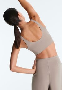 Žena s tmavým copom, ktorá sa natiahne do strany, oblečená v béžovej športovej podprsenke a legínach na jednoduchom svetlom pozadí.