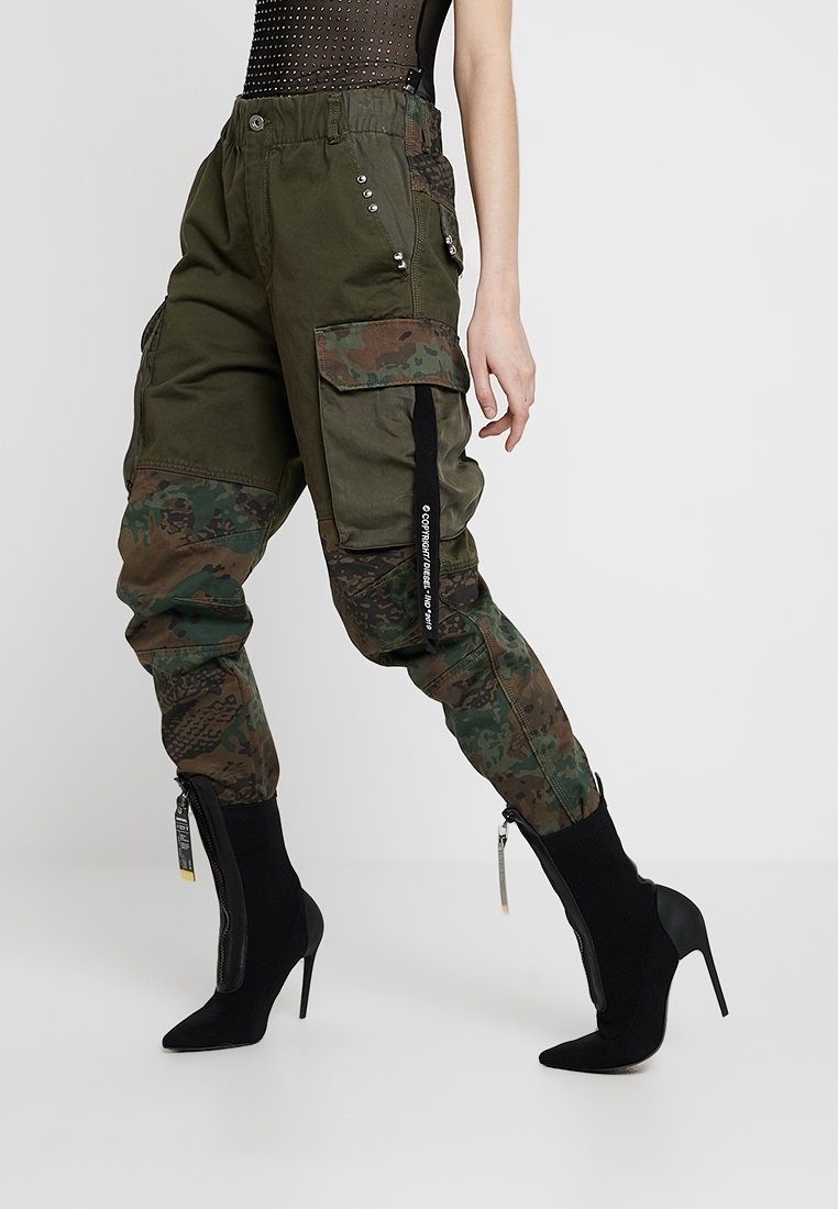 Personne portant un pantalon cargo camouflage vert et des bottines noires à talons hauts avec fermetures éclair à l'avant, debout devant un fond uni.