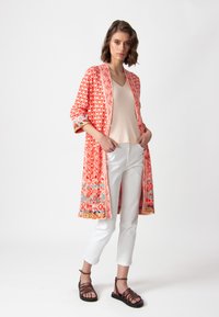 Garšs, atvērta priekšpuse kimono korālā krāsā ar ģeometriskiem rakstiem, trīs ceturtdaļu piedurknēm un krāsainu apdari, apvienots ar bēša kreklu un baltām biksēm.