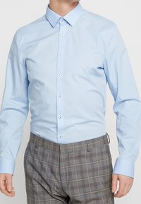 Lichtblauw overhemd met knopen, een middelgrote spreidkraag, lange mouwen en witte knopen. Draagt een grijze ruitbroek met een gestructureerde stof.