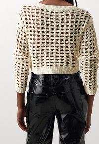 Sweter dziergany w kolorze kremowym, krótki, o otwartym wzorze splotu, z szerokim dekoltem i rękawami o długości 3/4, w zestawie z czarnymi błyszczącymi spodniami.