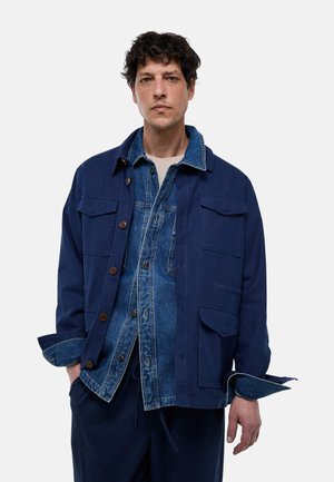 Homme portant des vestes bleues superposées, une en denim et une bleu marine, les mains dans les poches sur un fond blanc uni.