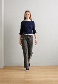 Pull marine, turtleneck blanc, jean gris et baskets noires. Le modèle se tient droit contre un mur gris uni sur un sol en bois.