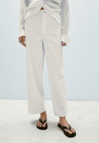 Hvide bukser med bred benvidde og en glat overflade samt knaplukning foran. Sammen med sorte flip-flops. Neutral baggrund.
