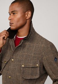 Bruine geruite wollen jas met een corduroy kraag, voorzien van twee voorzakken en messing knopen. Model past de kraag aan en draagt een rode trui.