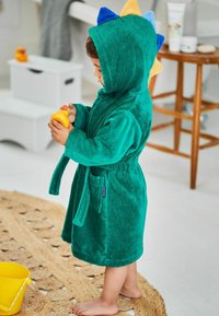 Zelený osuškový župan s kapucňou v tvare dinosaura, s modrými a žltými hrotmi, zviazaným pásom a bočnou kapsou. Dieťa držiace žltú gumenú kačicu.