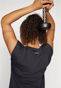 Czarny sportowy top bez rękawów z teksturowanym wykończeniem i luźnym krojem. Posiada małe logo i jest noszony podczas podnoszenia hantla.