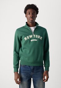 Sweatshirt zippé vert avec "NEW YORK" et "SCHOTT" imprimés en crème, associé à un jean bleu, avec des poignets et un ourlet côtelés.