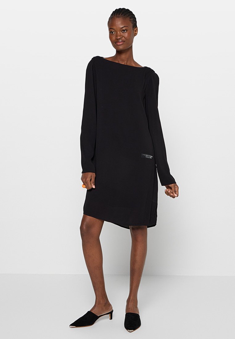 Abito nero a maniche lunghe con scollatura quadrata e apertura laterale. Tessuto liscio, lunghezza al ginocchio, con un sottile accento con logo vicino all'orlo.