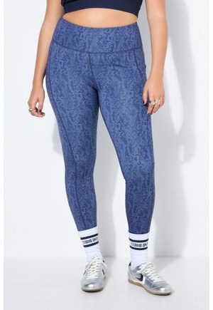 Person iført højtaljede, blå leggings med slangeprint, hvide sokker med sorte striber og tekst, samt sølvfarvede Nike-sneakers, stående mod hvid baggrund.