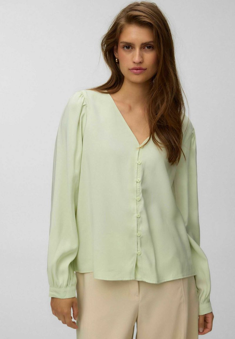 Jeune femme aux longs cheveux bruns, portant un chemisier vert pâle boutonné et un pantalon beige clair, debout sur un fond gris clair.