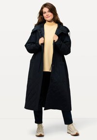 Lång mörkblå vadderad kappa med huva, med stora framfickor, knäppning fram och matchad med en ljusgul topp och sneakers.