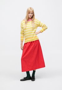 Farebný pletený sveter s kvetinovým vzorom v žltej, ružovej a bielej farbe na pruhovanom podklade. Dopĺňa ho dlhá červená plisovaná sukňa.