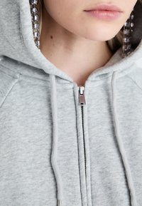 Šviesiai pilkas užtrauktukas su gobtuvu, pagamintas iš minkštos medžiagos, su gobtuvu, priekiniais užtrauktukais ir dekoratyviniais grandinėlės akcentais palei vidinius gobtuvo kraštus.