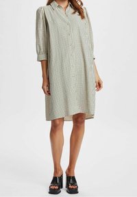 Abito camicia beige con motivo a quadri neri, maniche a tre quarti e vestibilità rilassata, abbinato a sandali platform neri.