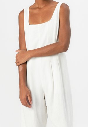 Combinaison en lin blanc avec un décolleté carré, des bretelles larges et un design plissé. Le tissu semble doux et léger.