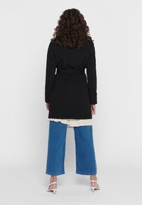 Dlouhý černý kabát s páskem, který má hladkou texturu. Kombinovaný s volnými modrými denimovými kalhotami a bílými sandály na podpatku. Pozadí je jednoduché bílé.