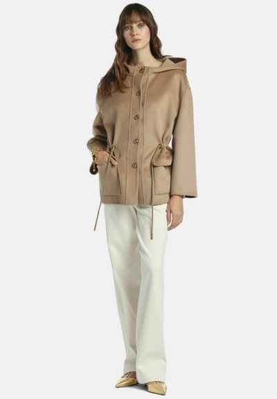 Jachetă bej cu glugă, nasturi aurii, talie cu șnur și două buzunare frontale, asortată cu pantaloni largi albi și pantofi aurii.