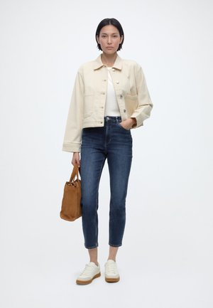 Kvinde står iført en lysebeige jakke, hvid top, mørkeblå skinny jeans, hvide sneakers og holder en brun håndtaske i venstre hånd.