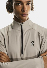 Šviesiai smėlinis sportinis ketvirčio užtrauktukų viršus su juodu užtrauktuku ir logotipu. Yra tekstūruota medžiaga, aukšta apykaklė ir prigludusi forma.