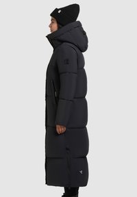 Donna che indossa un lungo cappotto invernale imbottito nero con cappuccio e un berretto di maglia nero, in piedi di profilo contro uno sfondo chiaro e uniforme.