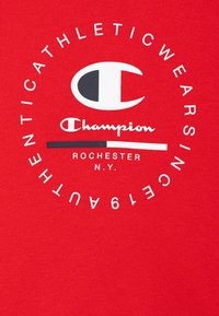 Camiseta de algodón roja con un gran logo circular que presenta una "C" estilizada en blanco y negro, y el texto "Champion Rochester N.Y." en blanco.