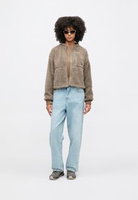 Žena s kučeravými vlasmi v béžovej fleecovej bunde na zips, svetlomodrých džínsach, sivých teniskách a slnečných okuliaroch v tvare mačacích očí stojaca na bielom pozadí.