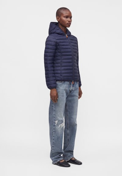 Personne portant une veste matelassée à capuche bleu marine, un jean ample usé et des chaussures noires sans lacets, debout devant un fond uni.