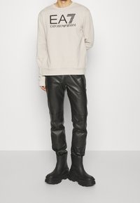 Světlé béžové tričko s černým logem EA7 Emporio Armani, doplněné o černé kožené kalhoty a robustní černé kotníkové boty. Jednobarevné a jednolité textury.