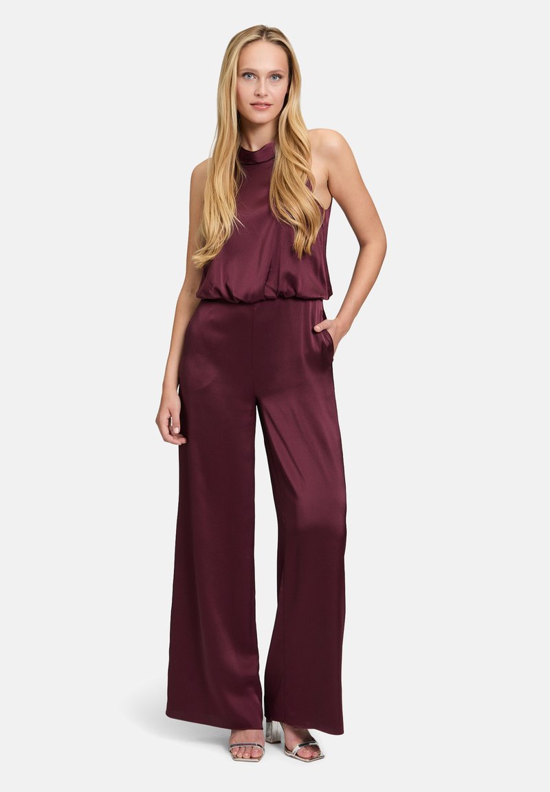 Burgundy jumpsuit with a halter neckline, wide legs, and a gathered waist. Made of smooth, shiny fabric with side pockets. Silver sandals featured.