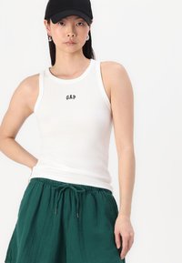 Weißes geripptes Tanktop mit kleinem schwarzen "GAP"-Logo, kombiniert mit lockeren grünen Shorts, vor einem schlichten Hintergrund.