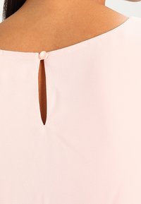 Blouse rose clair avec une petite ouverture en forme de clé au dos, fermée par un bouton circulaire. Tissu lisse avec une texture douce.