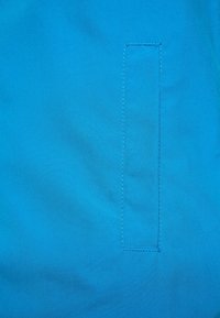 Jasnoniebieska tkanina o gładkiej fakturze posiada pionowy kontur kieszeni przeszyty nićmi. Wzdłuż krawędzi widoczna jest pojedyncza kontrastowa nitka.