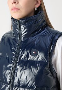 Gilet imbottito blu navy con colletto alto, texture lucida, chiusura con zip e patch con il marchio sul petto. Presenta cuciture orizzontali.