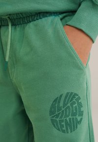 Žalios medvilnės kelnes su elastinga juosmena ir rankogaliais. Ypatybė – didelis, apvalus logotipas, spausdintas tamsesne žalia spalva ant šlaunies.