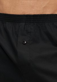 Taljebånd og øvre del af sorte boxershorts med knaplukning, båret af en person med synlig hud og kropsbehåring over taljebåndet.