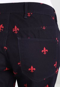 Tmavě modré kalhoty s opakujícím se červeným vzorem lilie, s viditelnou zadní kapsou a stehováním na pásku.