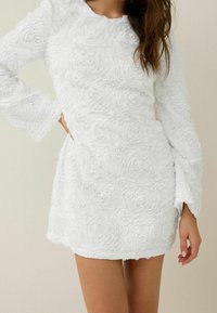 Robe blanche en tissu texturé avec des motifs de roses, dotée de manches longues et d'un col rond, se terminant au-dessus de la mi-cuisse.