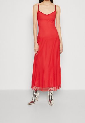 Vrouw draagt een mouwloze rode jurk met dunne bandjes en zilveren puntige hoge laarzen, staand tegen een effen witte achtergrond.