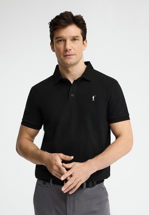 Mężczyzna noszący czarną koszulkę polo z krótkim rękawem z małym białym logo, szare spodnie, stoi na jasnym, jednolitym tle z rękami splecionymi.