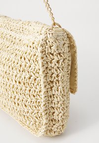 Bolso de crochet beige con cierre de solapa y una correa de cadena dorada sobre un fondo blanco.