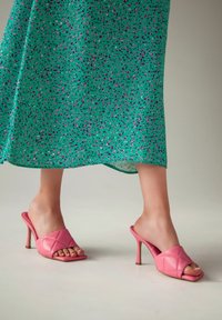 Mule roz cu toc, din piele matlasată, cu spatele deschis și vârf pătrat. Purtați cu o rochie verde cu model, având desene mici și dispersate.