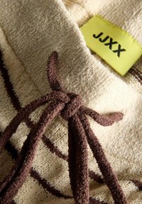 Tanka tkanina s smešnim prugama; ima vezivanje i svijetlu žutu oznaku s natpisom "JJXX." Mekana tekstura i uočljivi tkanini detalji.