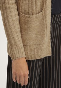 Beige rebrovaný pletený cardigan s prednou vreckom a priliehavými rukávmi, skombinovaný s čiernymi pásikovanými nohavicami. Mäkko textúra je evidentná.